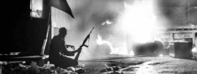 Joe McCann crouching with a rifle beside a burning barricade at Inglis’s bakery, Belfast, 10 August 1971. Photograph by Ciaran Donnelly